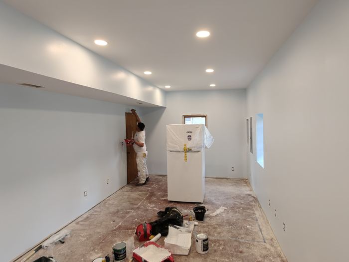 Painter working in a partially finished room with recessed lights and covered refrigerator.